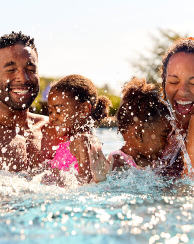 Family with two young children laughing and splashing together in a swimming pool on a sunny day.