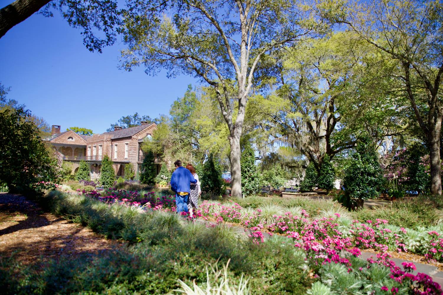 Visitors walking through a landscaped garden with blooming flowers and a historic brick home in the background
