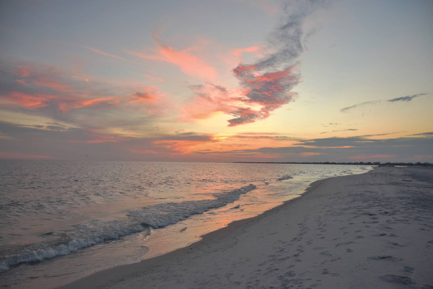 Sunset over a quiet sandy beach with gentle waves, footprints along the shoreline, and pink clouds reflected across the water