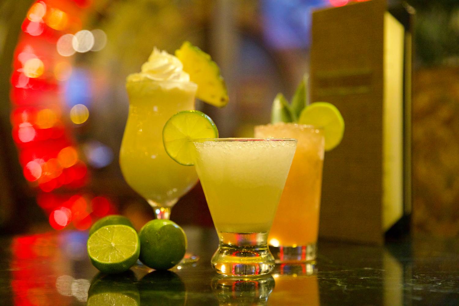 Three tropical cocktails garnished with lime and pineapple on a bar top with fresh limes and colorful lights in the background.