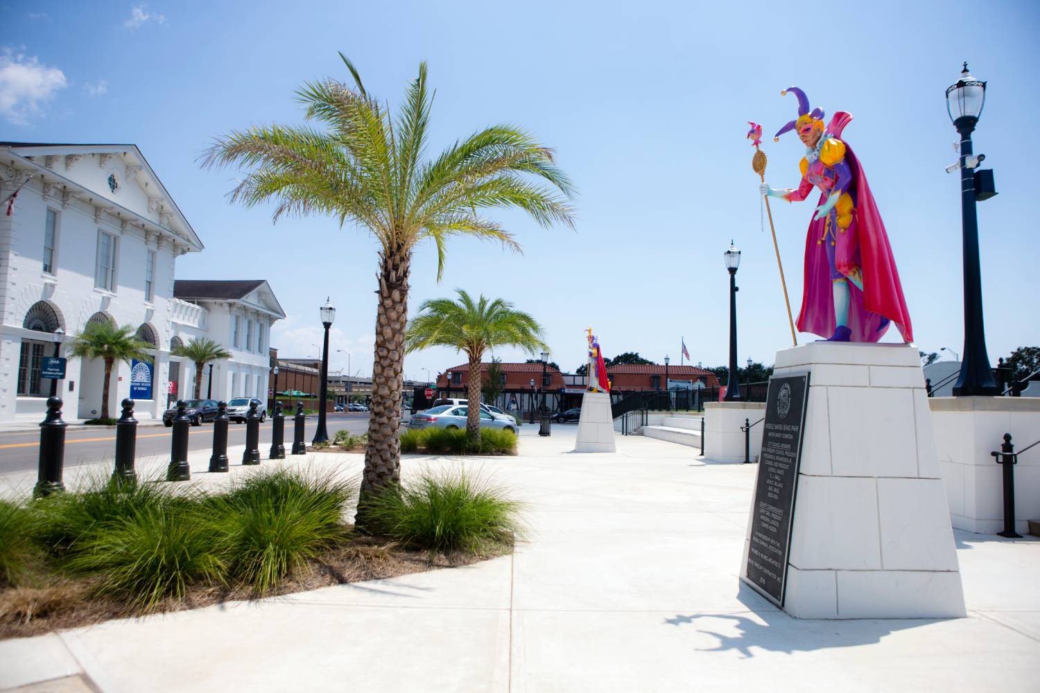 Colorful Mardi Gras jester statue displayed along a palm-lined waterfront promenade on a sunny day.