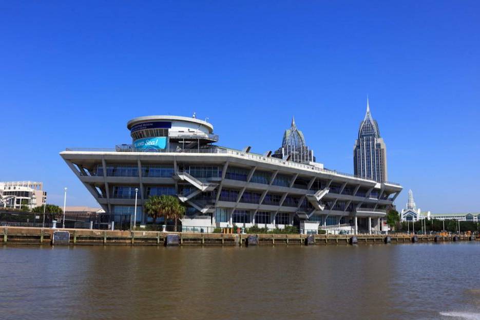 Mobile Convention Center on the waterfront with downtown Mobile skyscrapers rising behind the building under a clear blue sky.