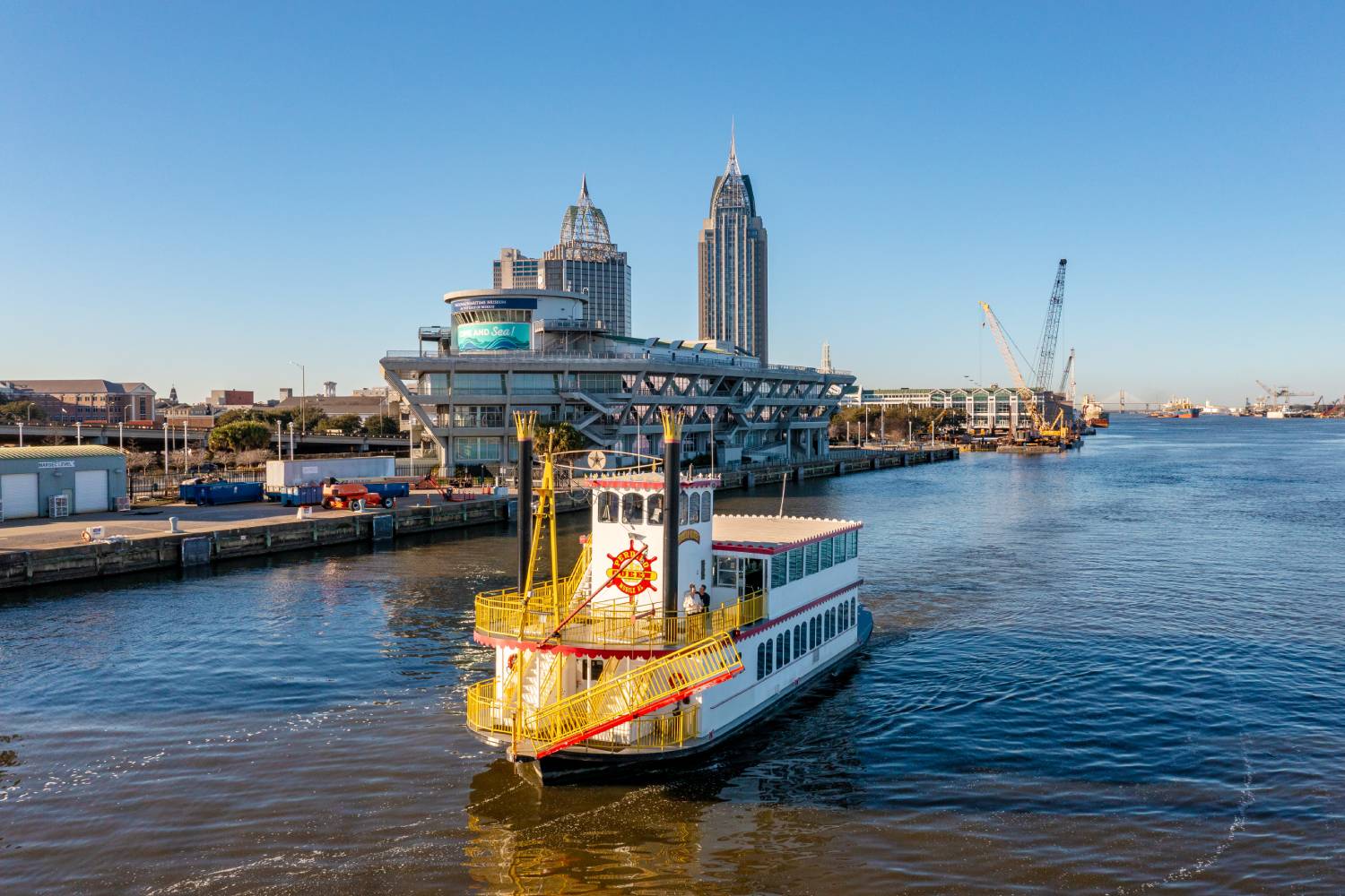 Riverboat cruise vessel traveling along Mobile River with the Mobile Convention Center and downtown skyline in the background.