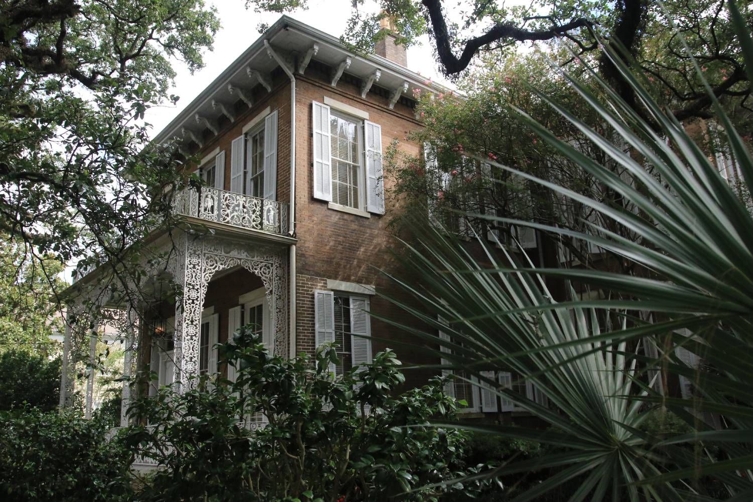 Historic brick home with ornate wrought-iron balcony and lush Southern gardens shaded by mature oak trees.