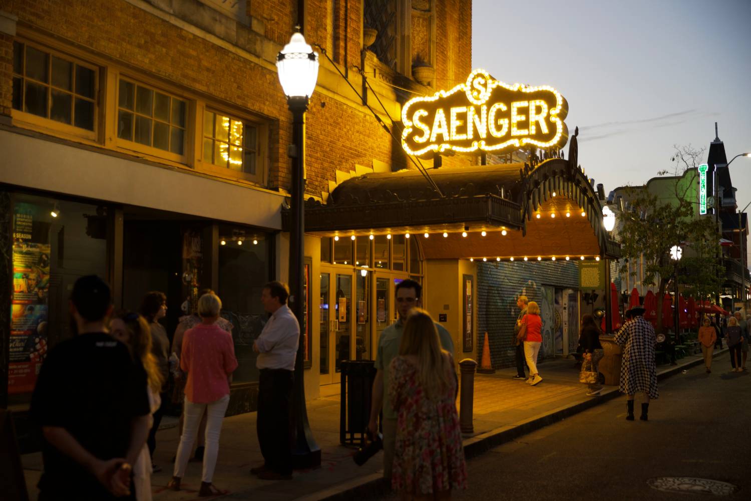 Crowds gather outside the illuminated Saenger Theatre marquee on a lively downtown street at dusk.