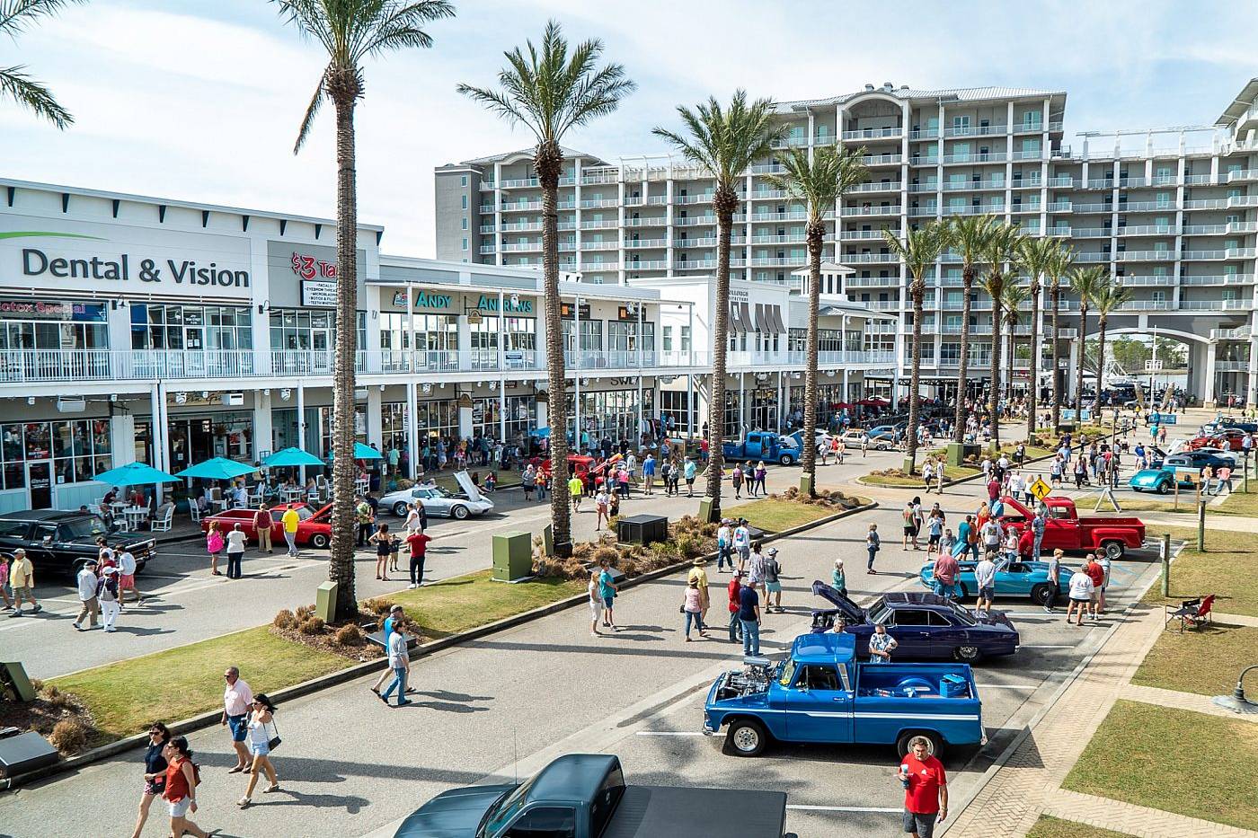 Crowds browse classic cars and trucks along a palm-lined boulevard in a busy waterfront entertainment district.