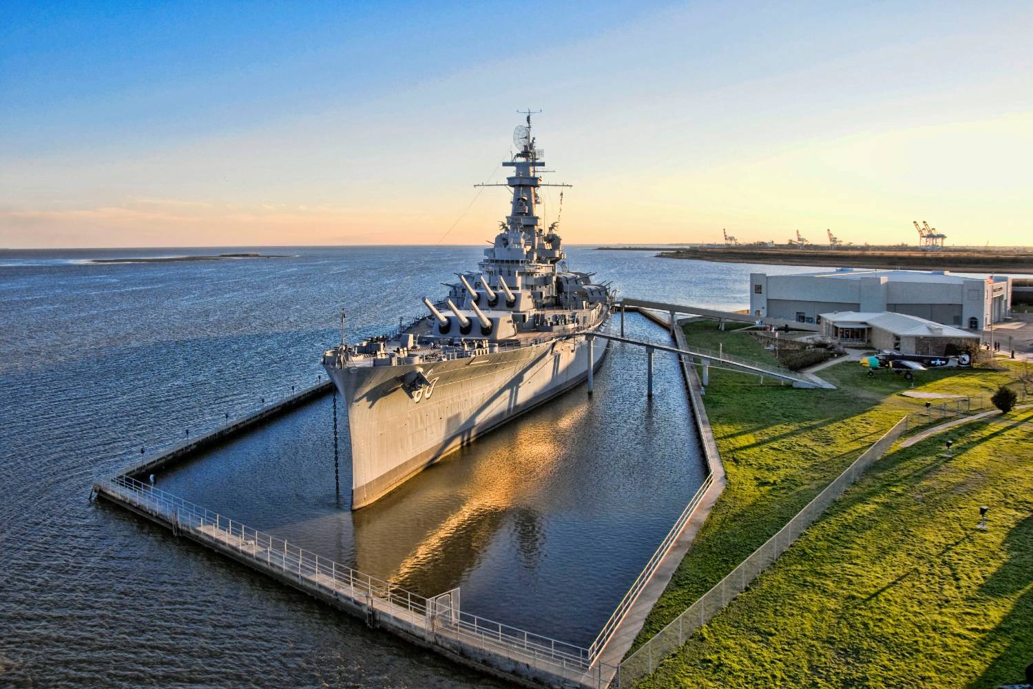 Battleship museum ship moored along the waterfront at sunset, viewed from above with harbor and shoreline in the background.